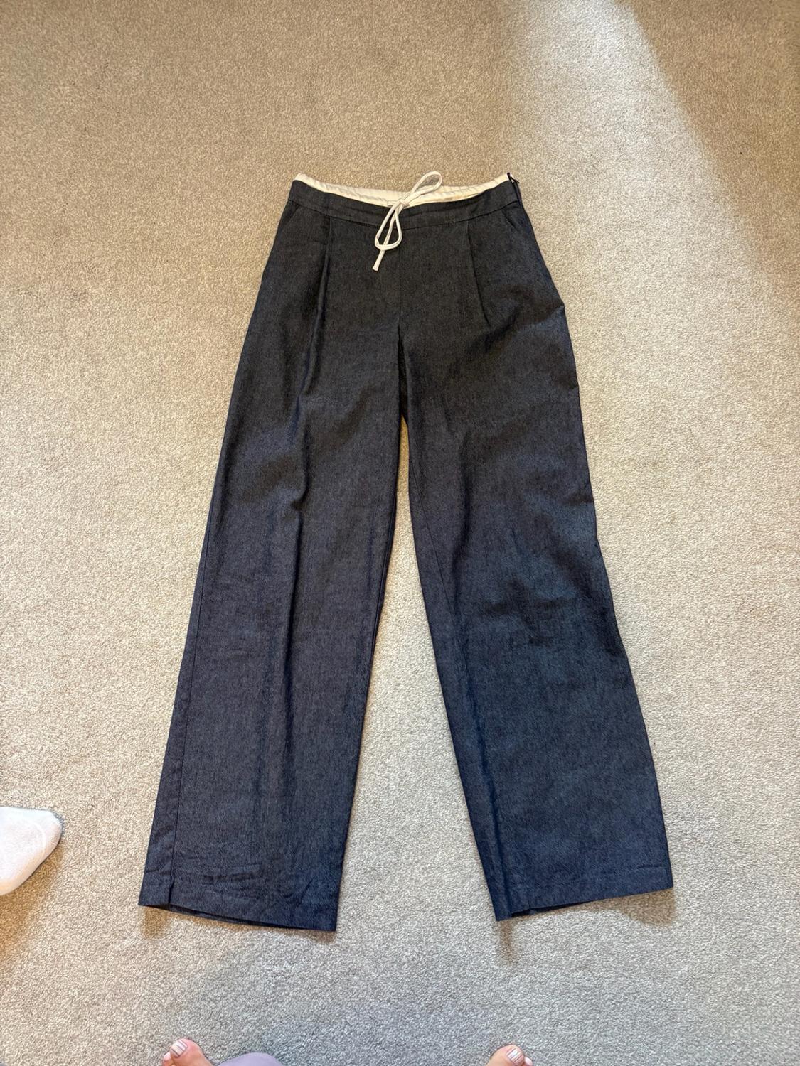 Dark grey wide-leg trousers with pleats and a white drawstring, laid out on a beige carpet. Bare feet are visible at the bottom edge.