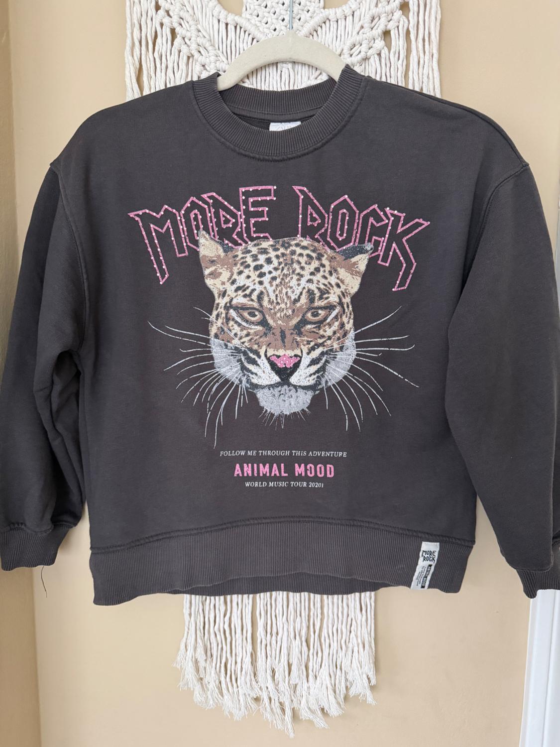 Black sweatshirt with leopard print design and "MORE ROCK", "ANIMAL MOOD" text, hanging on a hanger against a white macrame background.