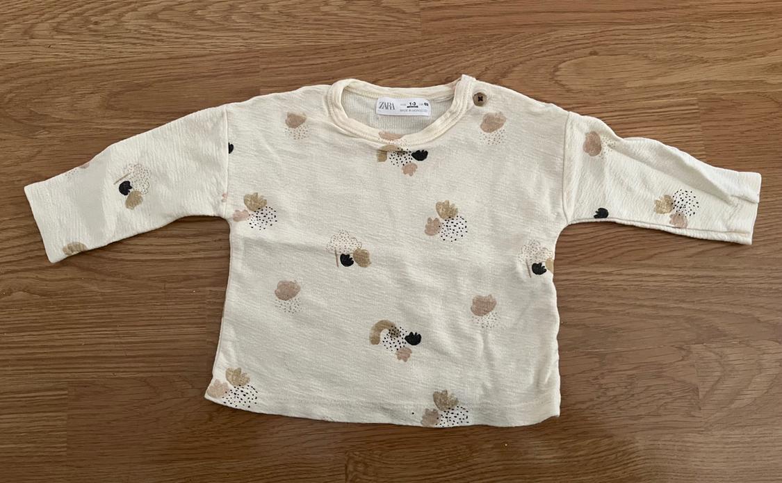Cream long-sleeved baby T-shirt with a floral print in beige and black tones, on a light wood background.