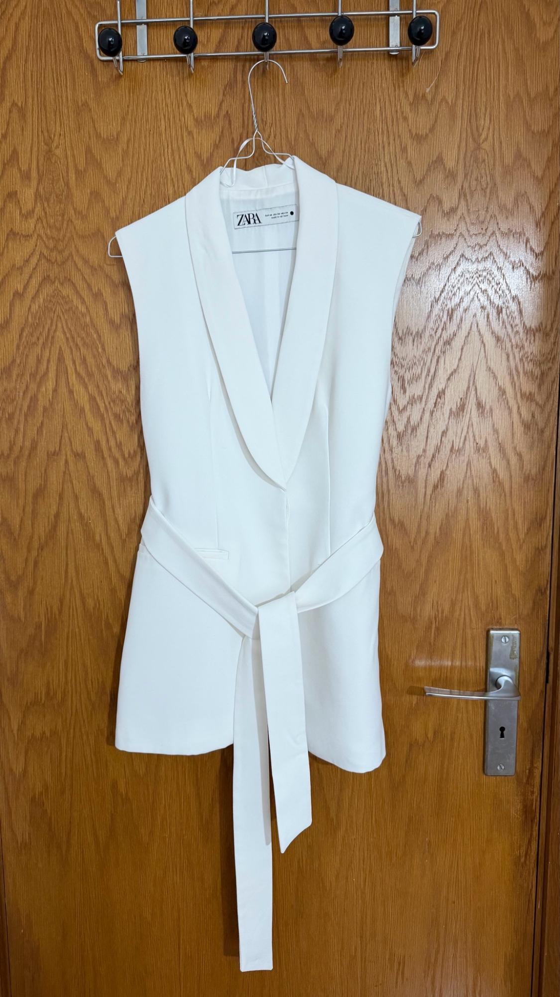 White blazer-style waistcoat with a tied belt, hanging on a coat hanger on a wooden door. ZARA can be read on the label.