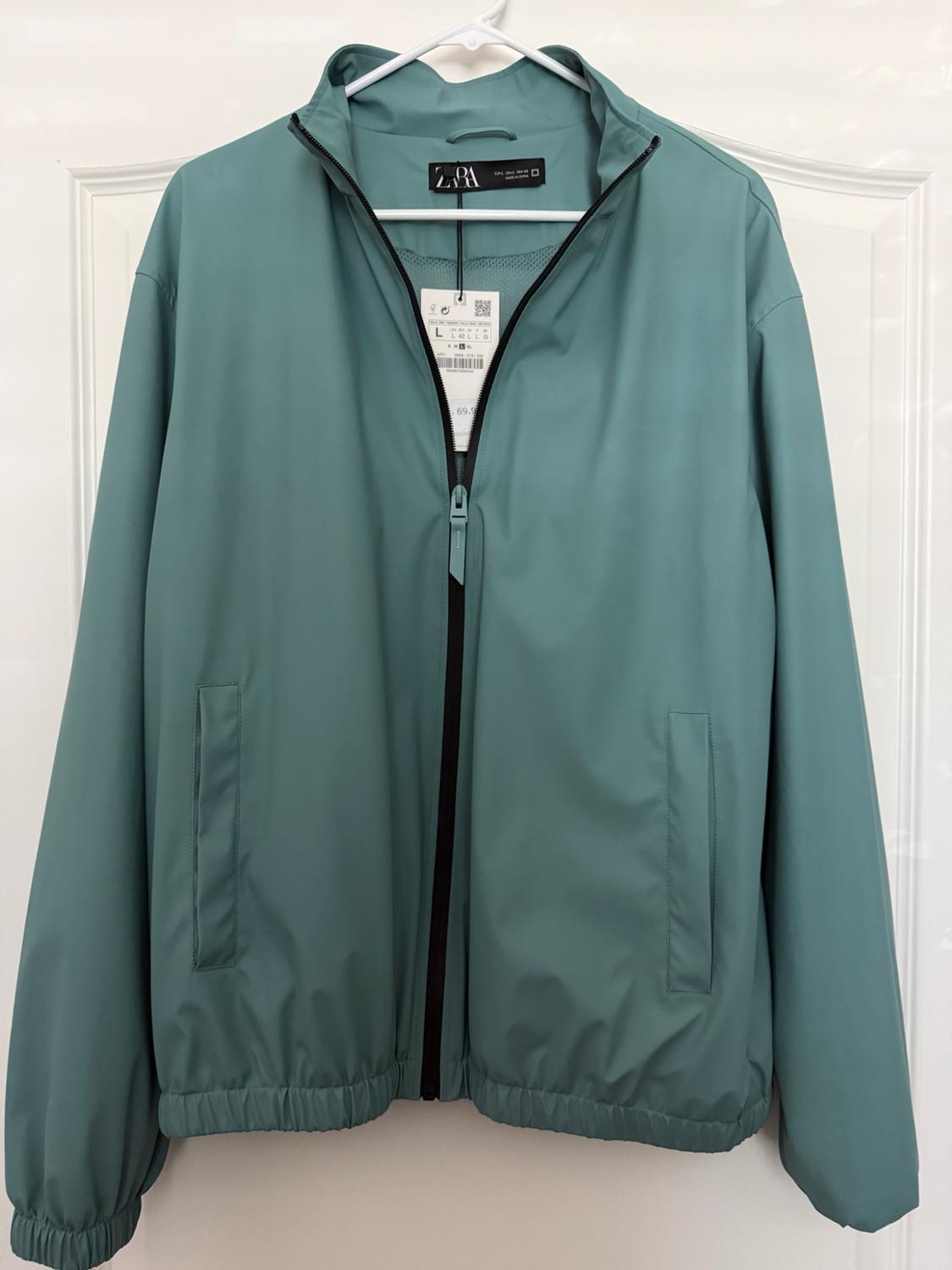 Green jacket with a black center zipper and pockets, hanging on a hanger over a white door. Text: ZARA, L.