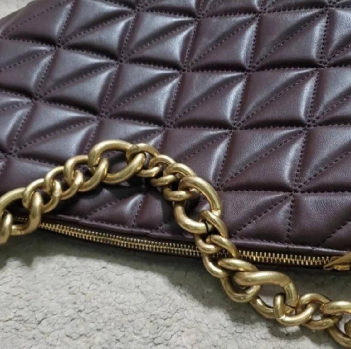 Close-up of a dark brown quilted handbag with a triangle pattern, zip, and chunky gold chain against a light background.