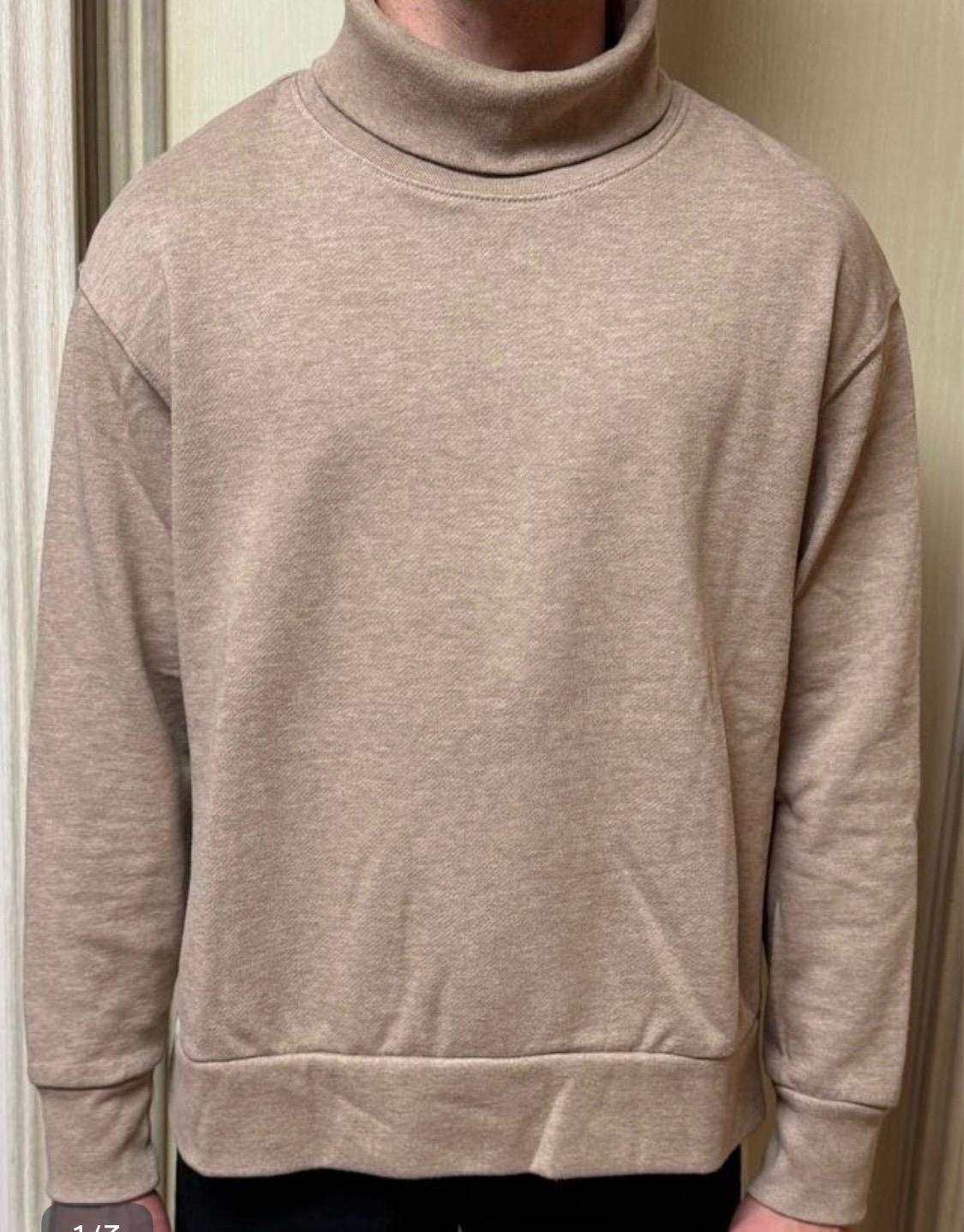 Model wearing a beige roll-neck jumper with long sleeves and a loose fit. Neutral light wood background.