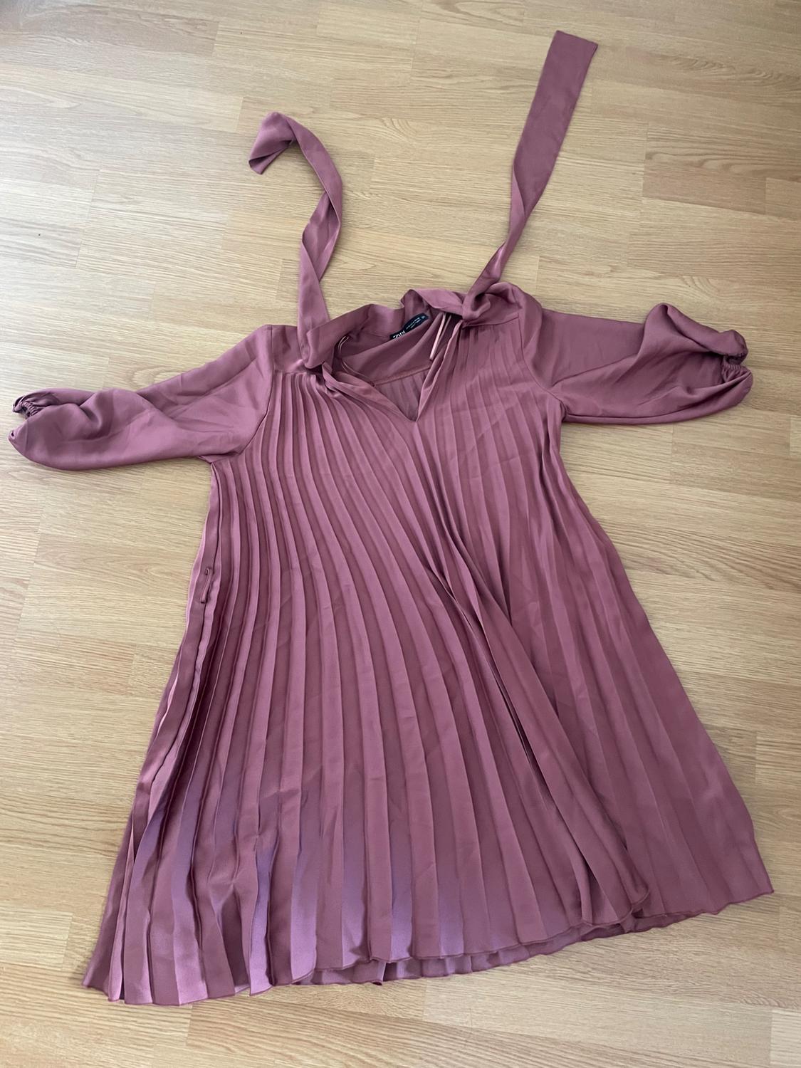 Pink pleated dress with a V-neck, ties and long sleeves with elasticated cuffs, laid out on a light wooden floor.
