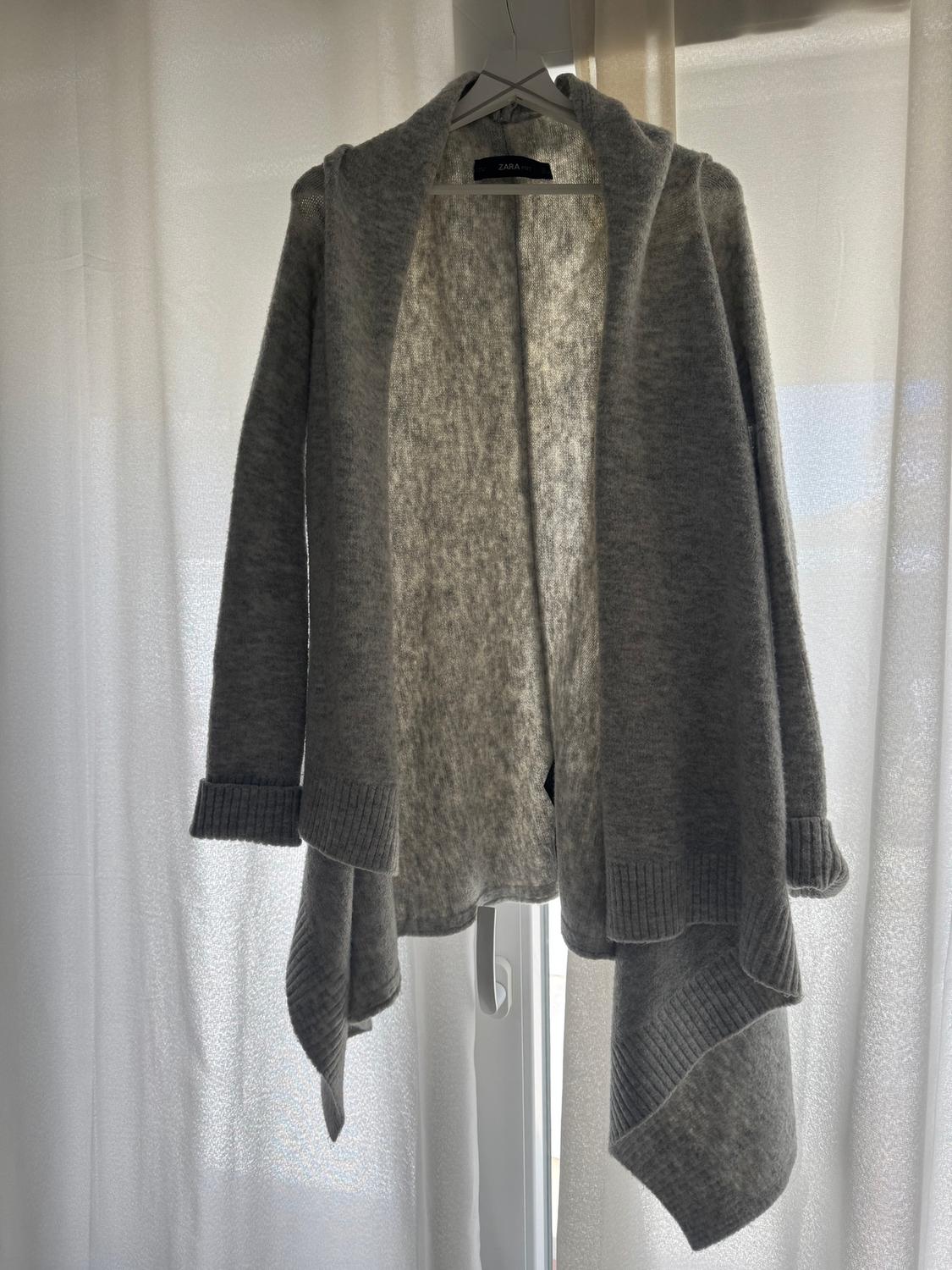 Grey wool cardigan hanging on a white hanger in front of light-coloured curtains. Visible label: "ZARA".