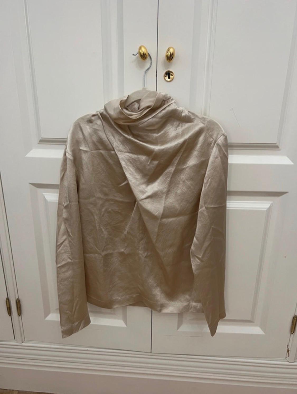 Light beige long-sleeved, high-neck satin blouse, hanging on a hanger against white double doors with gold knobs.