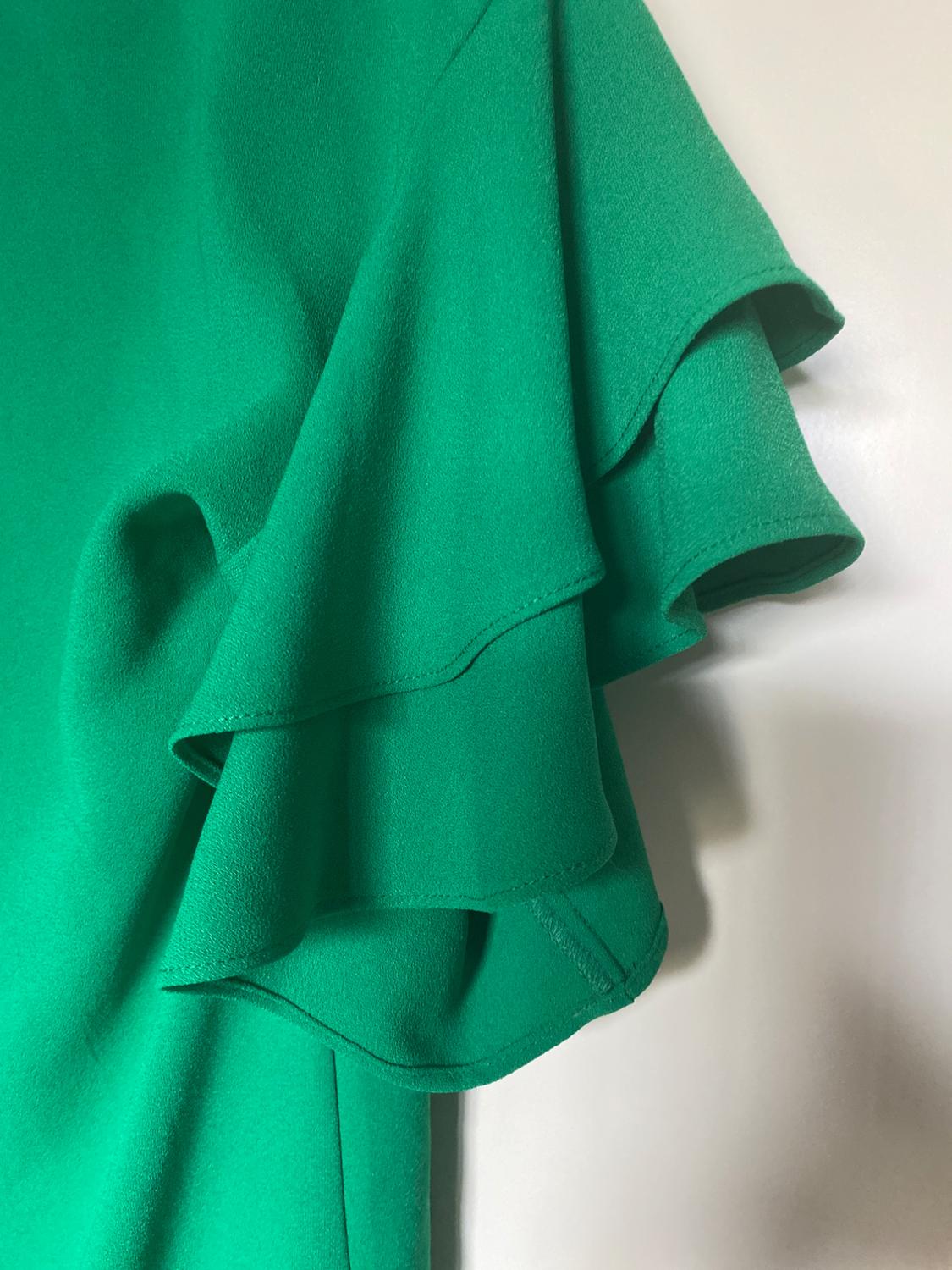Detail of a green blouse with ruffles on the sleeve, against a white background. The fabric has a soft and fluid texture.