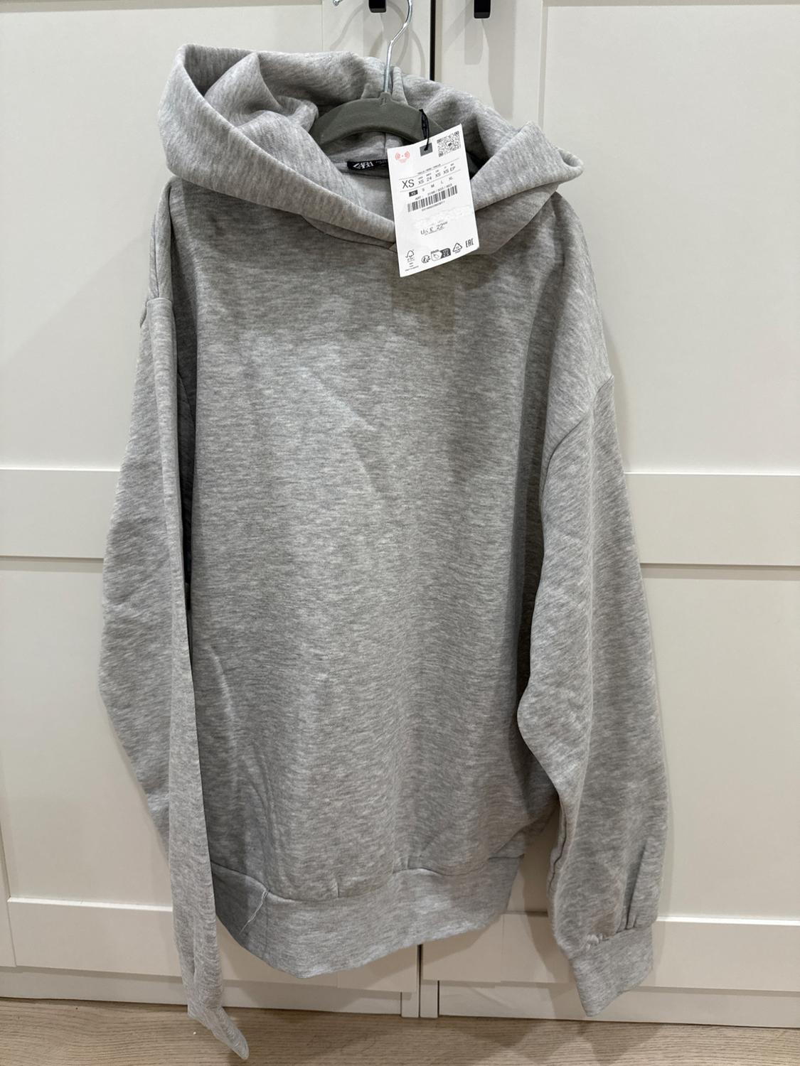 Grey hoodie hanging on a hanger, visible label with XS size details, on a white wardrobe background.
