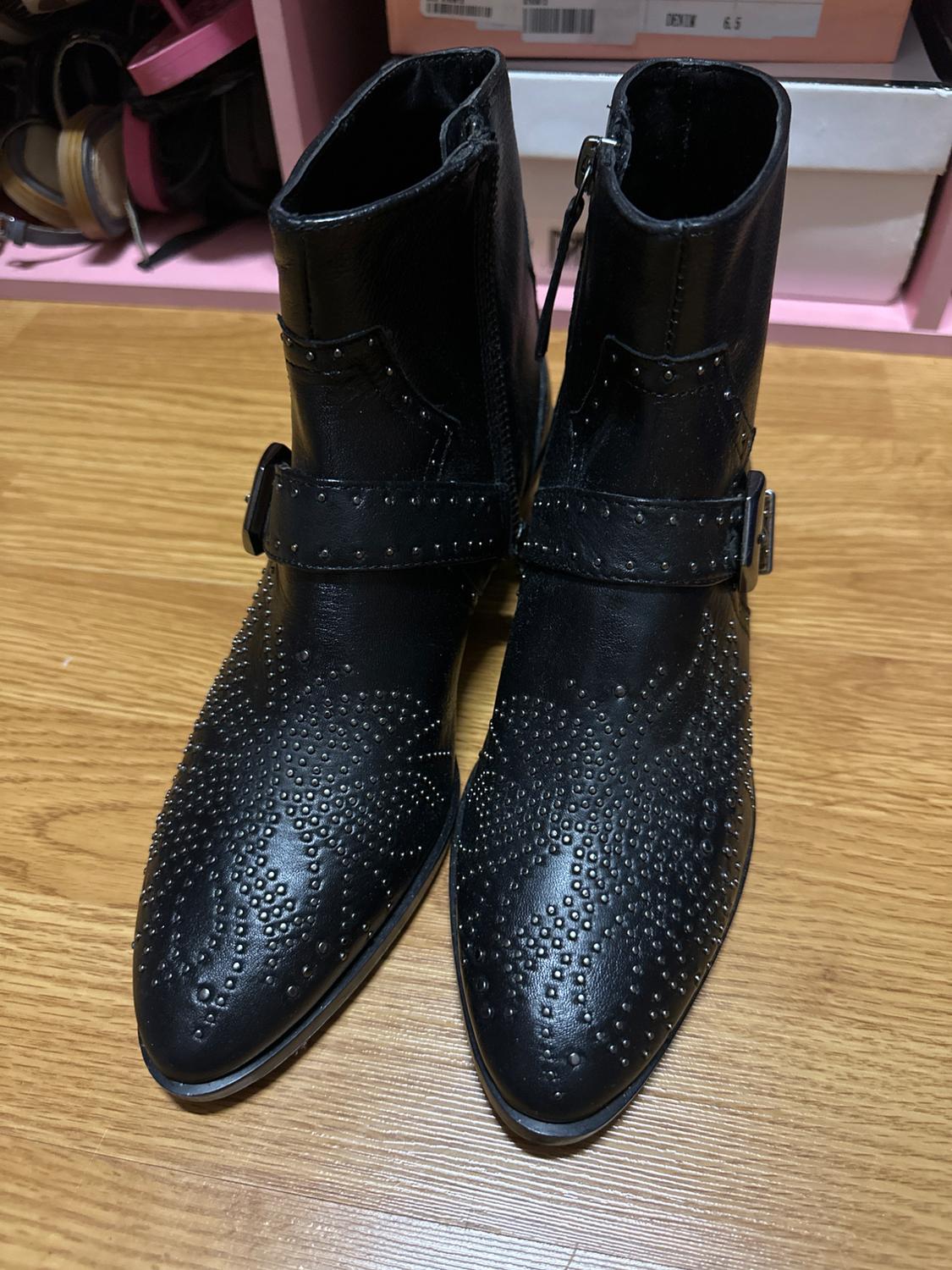Black ankle boots with decorative studs on the upper part, on a wooden floor. Background with boxes and ribbons.