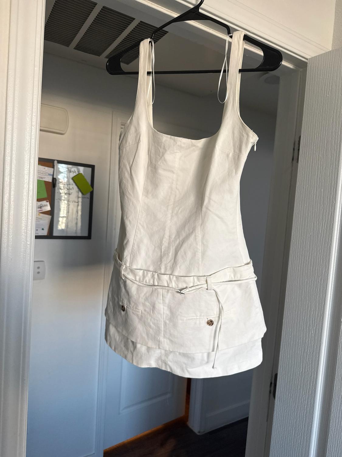White strap dress hanging on a door, with a square neckline, belt, and buttons. In the background, "Tasks" is written on a chalkboard.