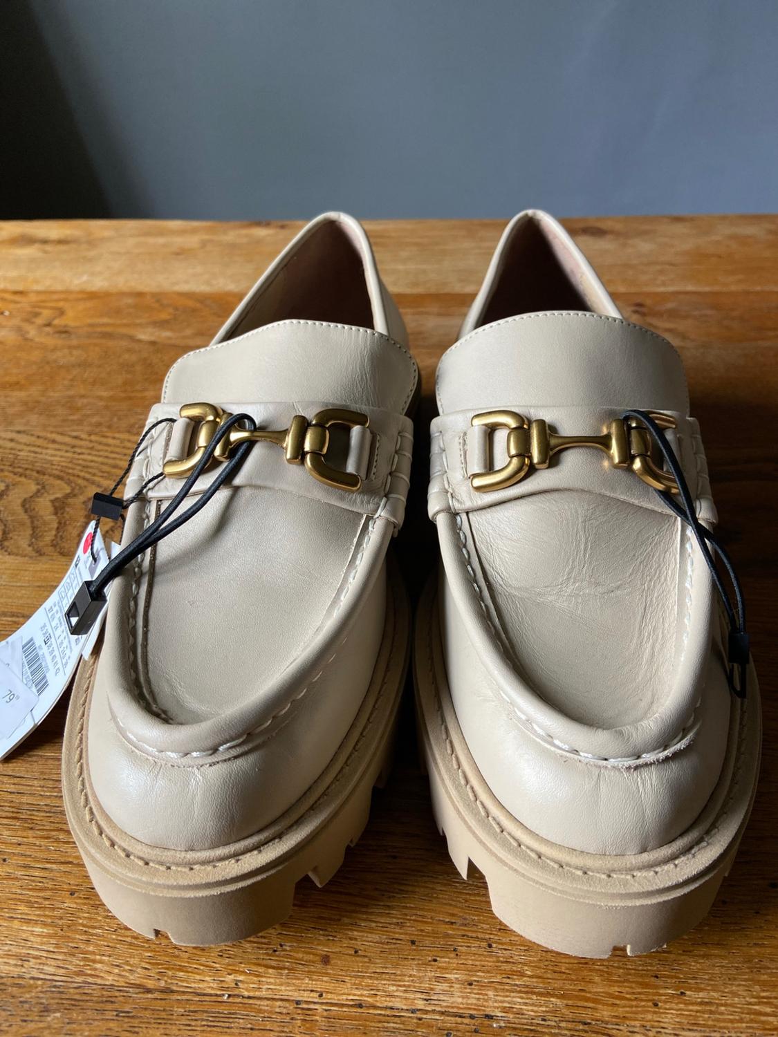 Pair of beige loafers with a chunky sole and gold metallic detail, resting on a wooden surface.