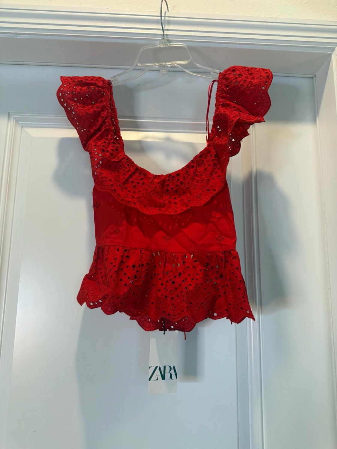 Red top with ruffles and eyelet embroidery hanging on a white door. Label with the text ZARA.