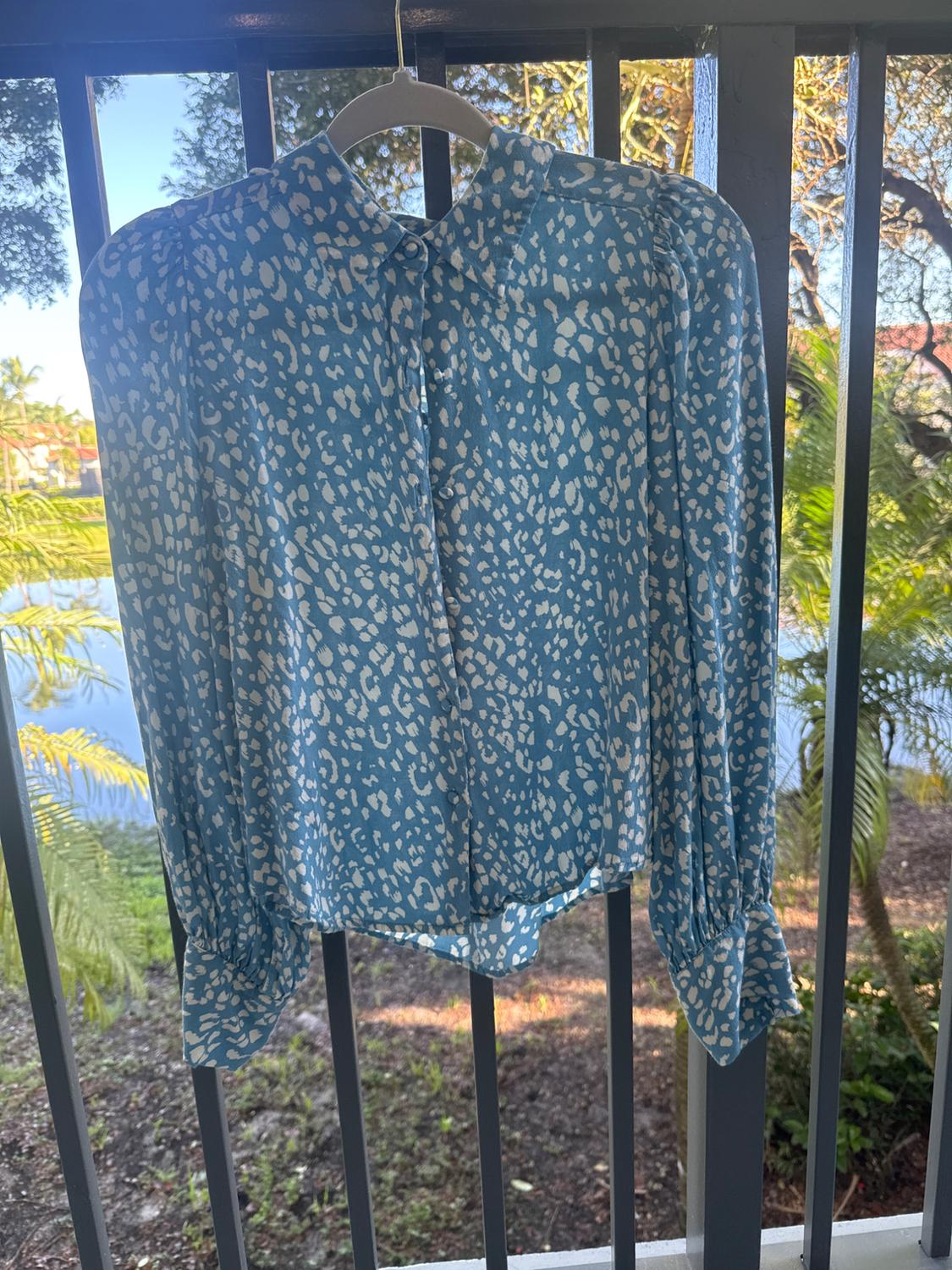 Blue blouse with white print hanging on a balcony, surrounded by greenery with a view of a lake and trees in the background.