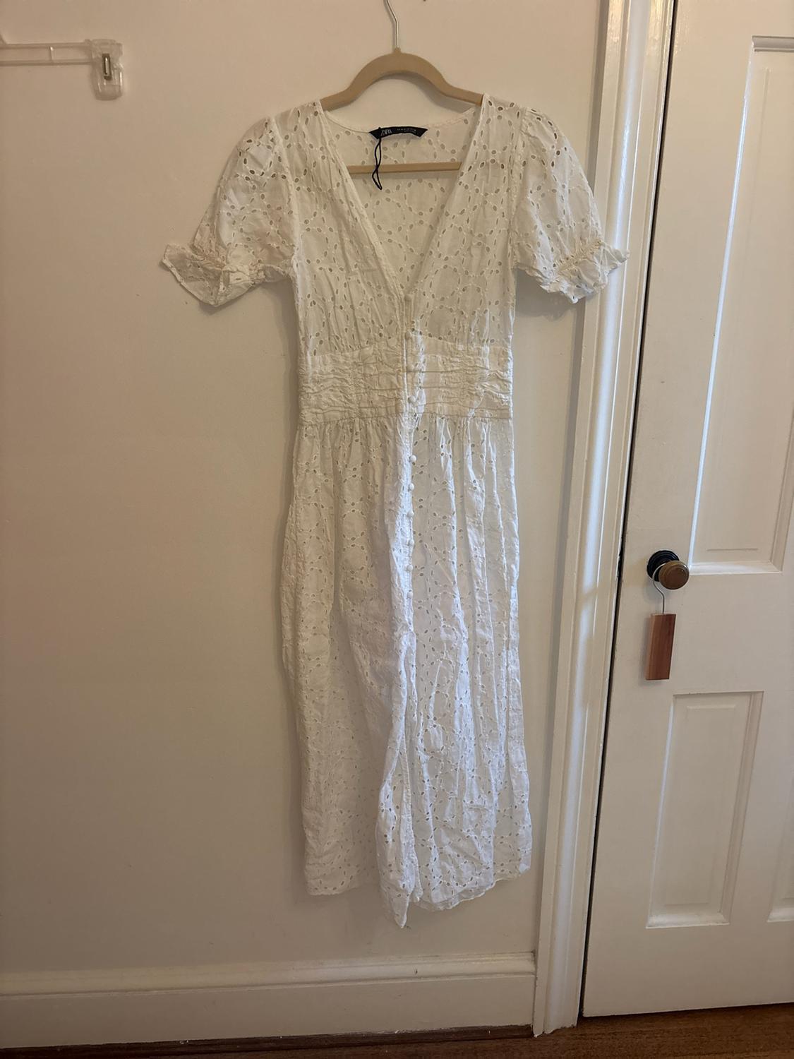 White dress with eyelet embroidery, short sleeves, V-neck, and front buttons, hanging on a hanger against a white wall.