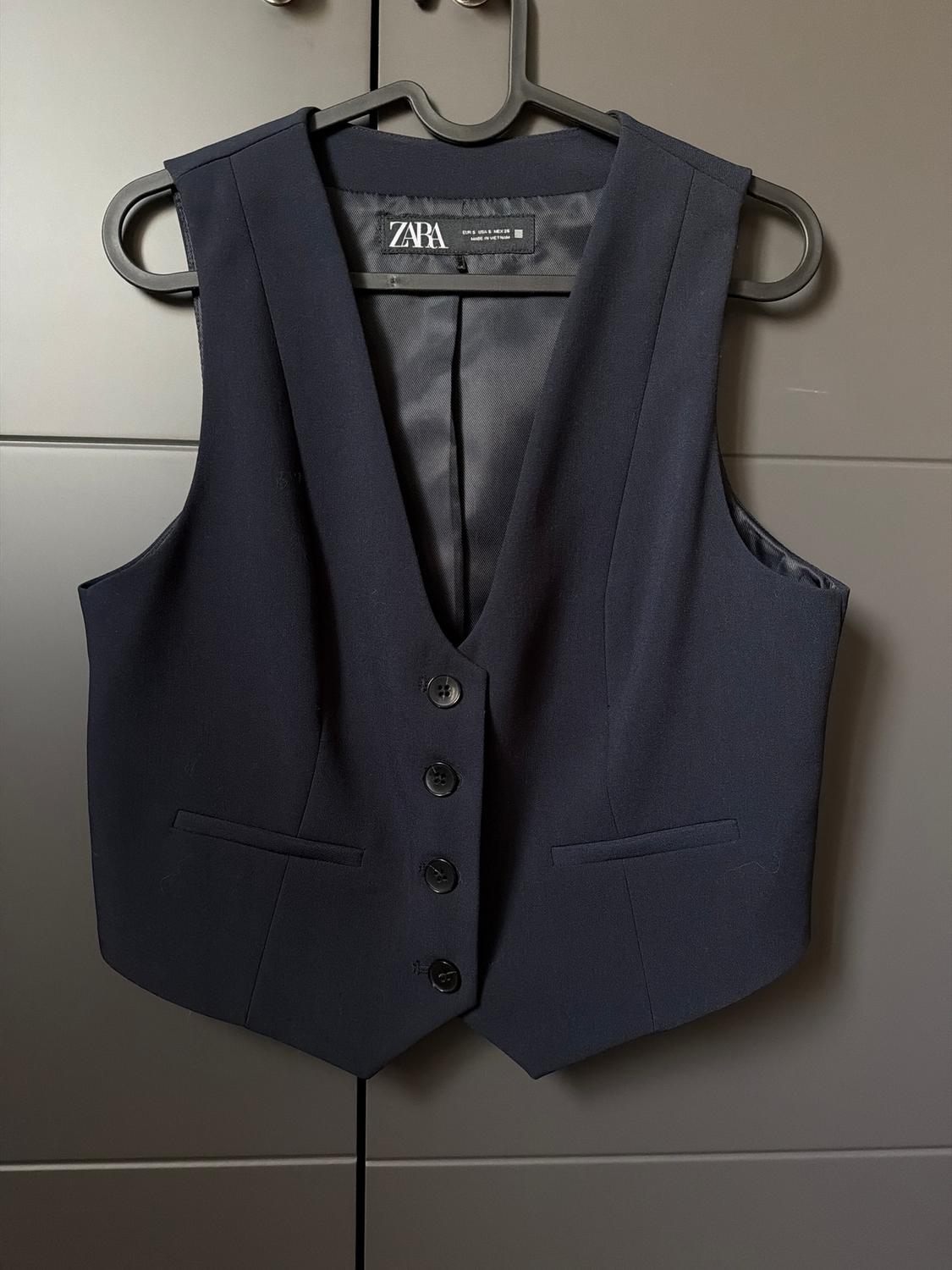Black dress waistcoat hanging on a black hanger, with five front buttons and a visible Zara label. Grey wardrobe background.