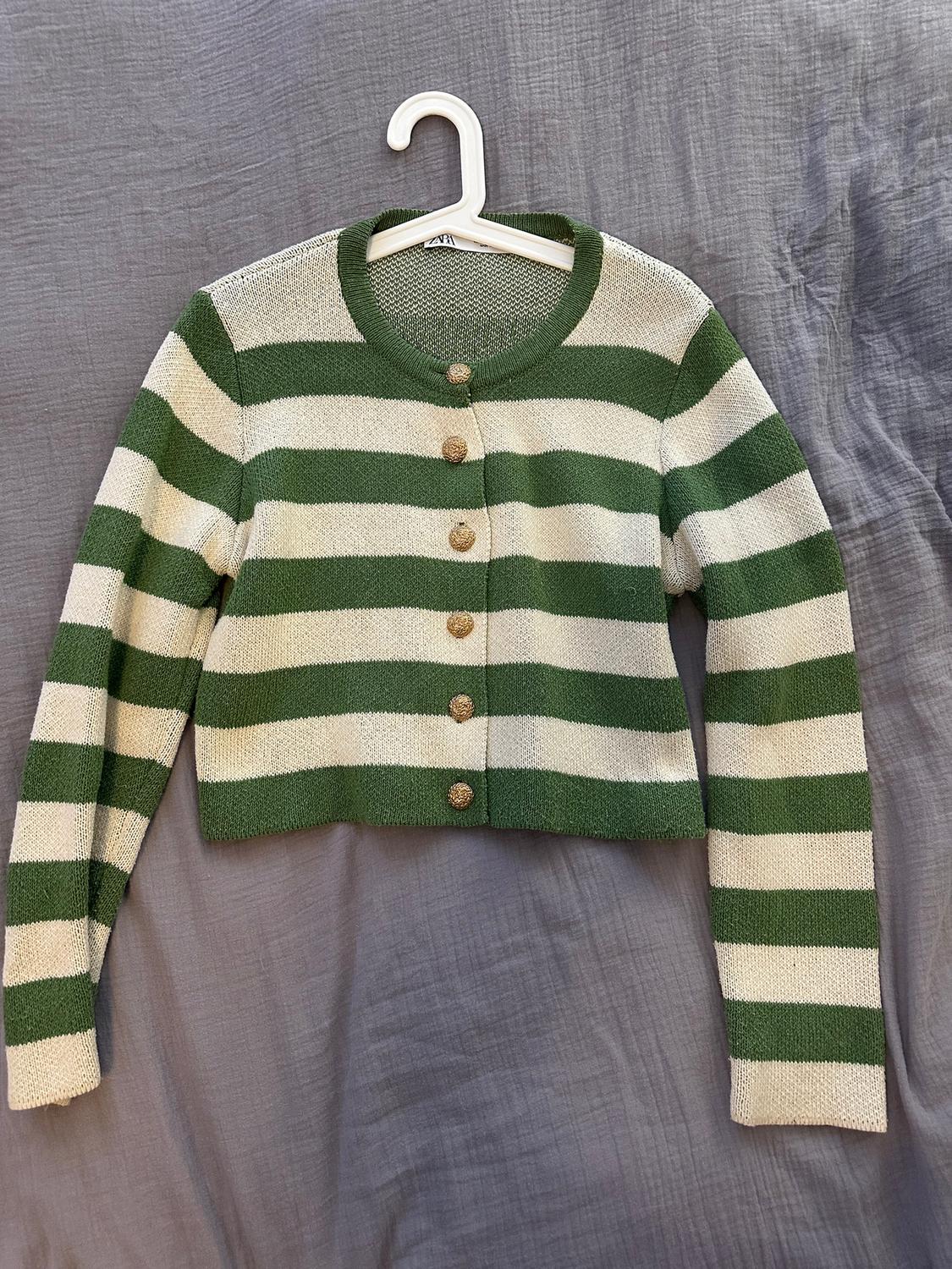 Green and white striped cardigan with gold buttons, hanging on a white hanger against a wrinkled grey background.