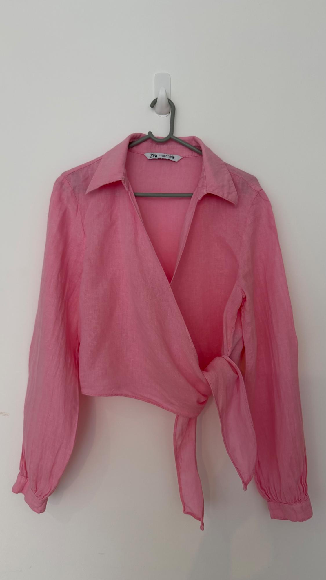 Pink long-sleeved wrap blouse with knot detail, hanging on a hanger against a white wall. Label with text ZARA.