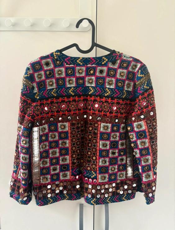 Colourful jacket with geometric patterns and sparkling details, hanging on a black hanger against a light background.