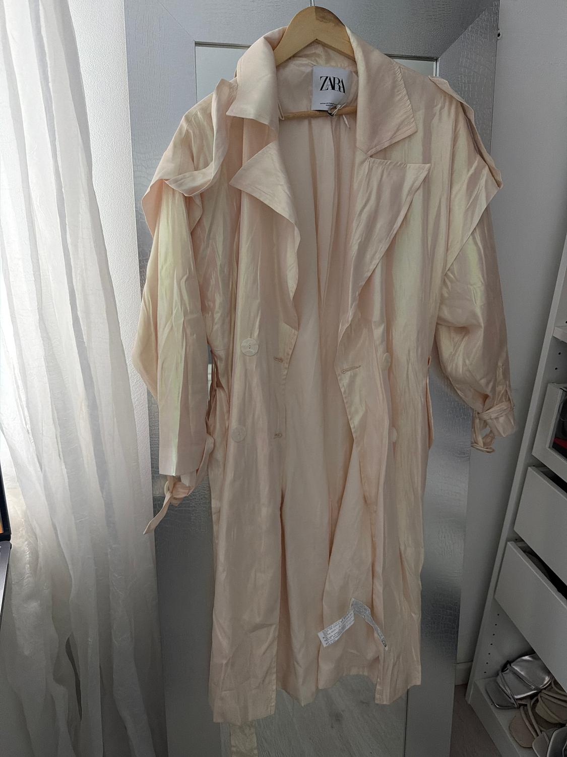 Beige coat hanging on a hanger with a Zara tag, next to a white curtain and a wardrobe with shoes in the background.