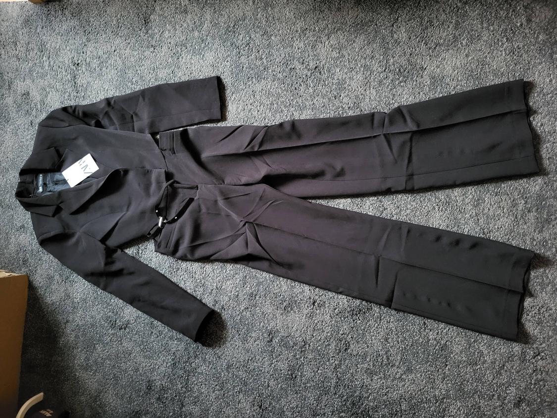 Two-piece black suit with jacket and trousers, label visible on grey carpet.