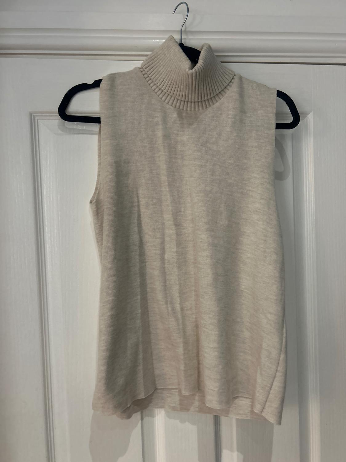 Beige sleeveless polo neck jumper, hanging on a black hanger over a white door.