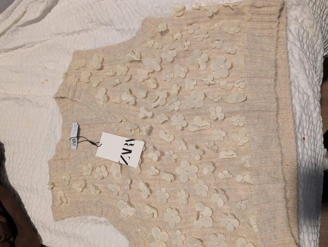 Beige vest with embossed floral appliqués on a wrinkled white surface. Label with "ZARA" text hanging from the vest.