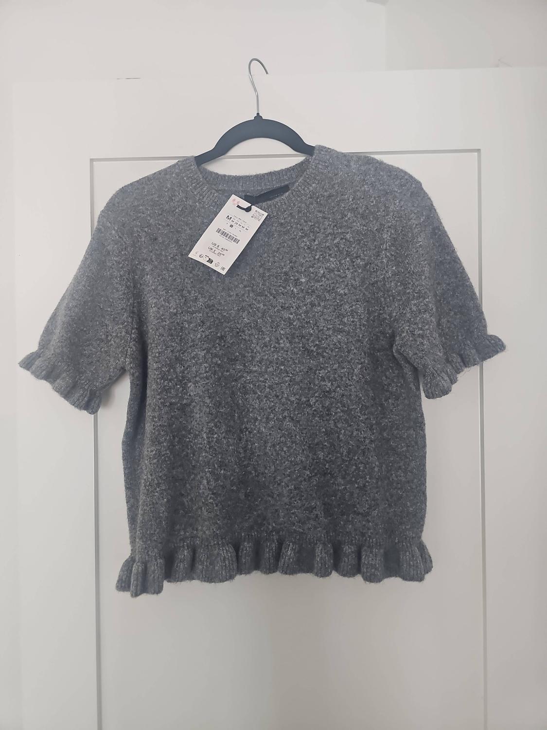 Grey short-sleeved knitted jumper with frills on the edges, hanging on a black hanger on a white door.