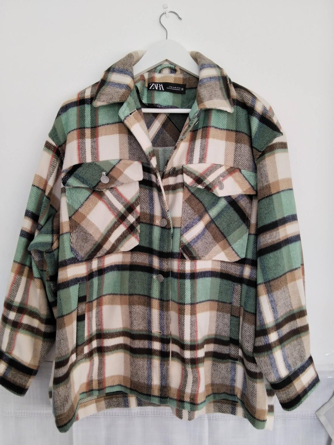 Checked overshirt in green, beige and black tones with front pockets, hanging on a white hanger. ZARA label visible.