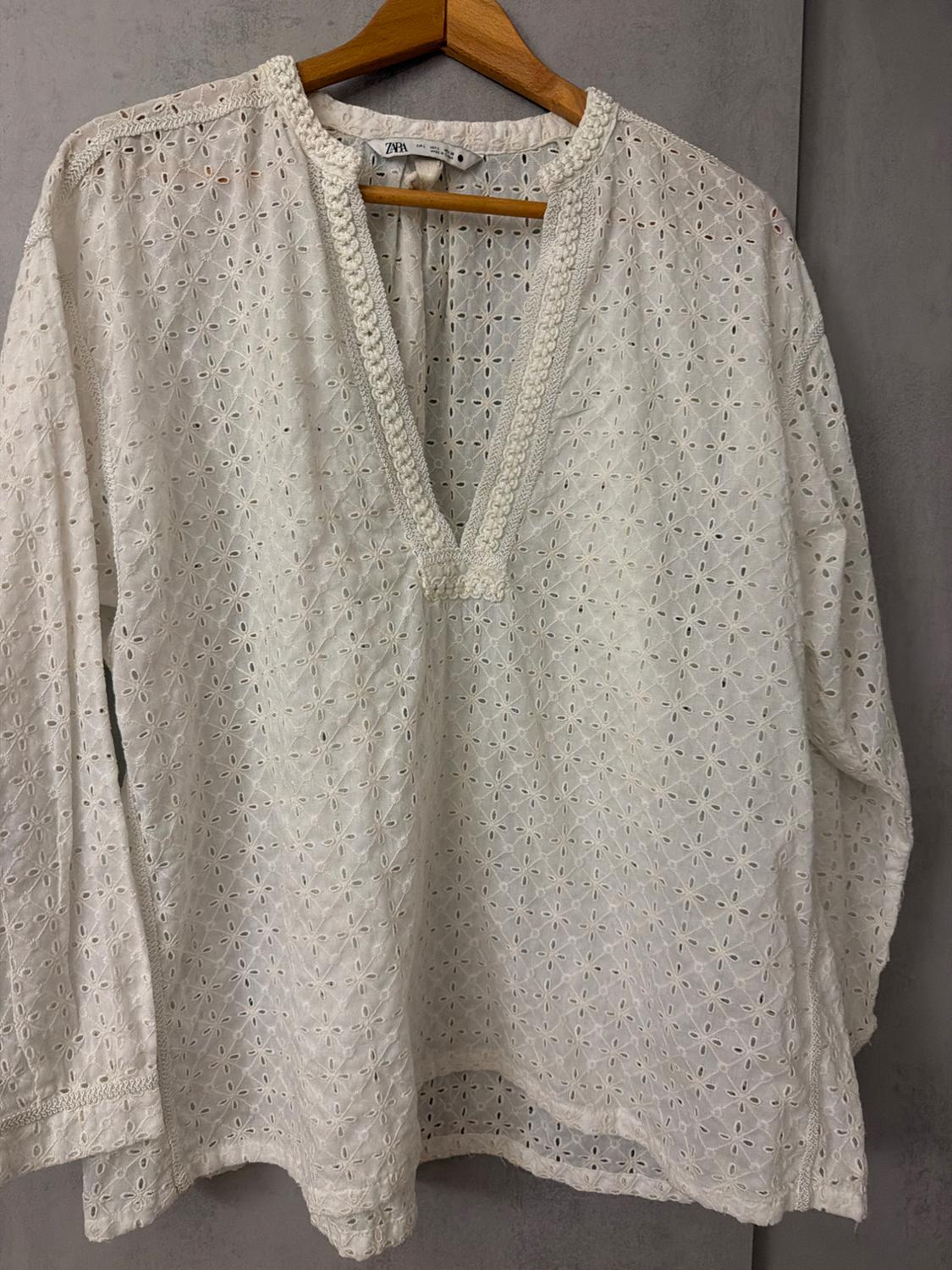 White blouse with floral eyelet embroidery, long sleeves and V-neck, hanging on a wooden hanger against a grey background. Text: ZARA.