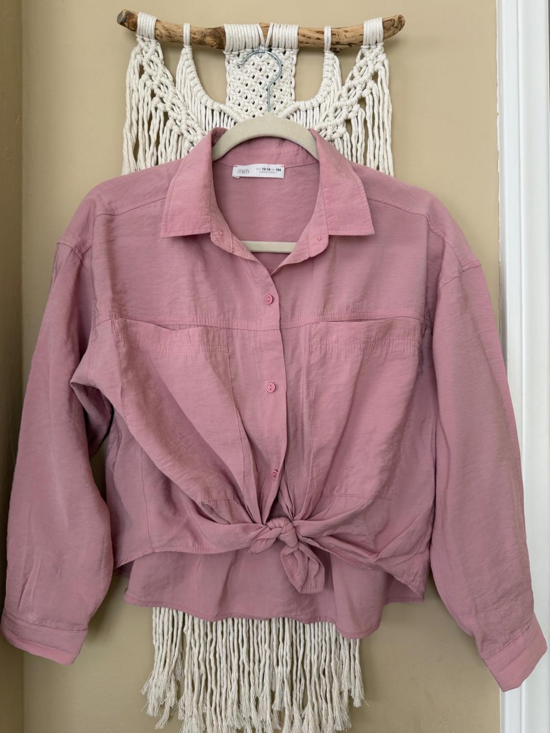 Pink long-sleeved shirt tied at the front, hanging on a macrame hanger against a beige wall. Visible tag: "Zara".