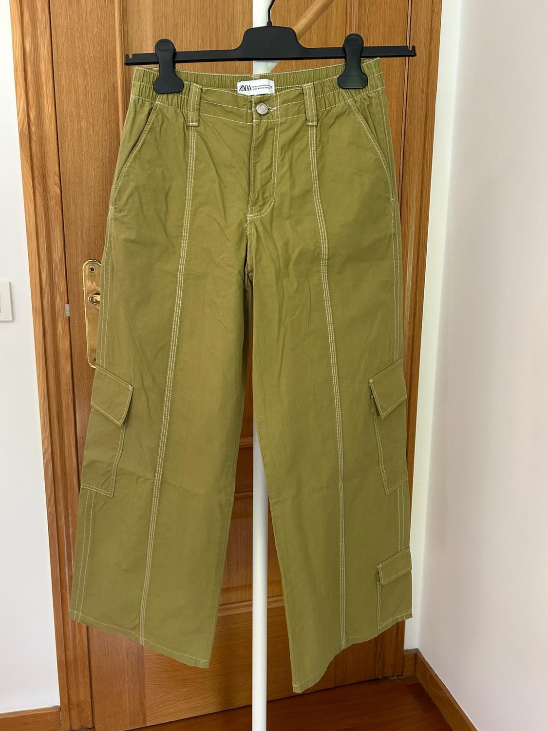 Olive green cargo trousers with white stitching and pockets, hanging from a clothes hanger in front of a wooden door. ZARA label visible.