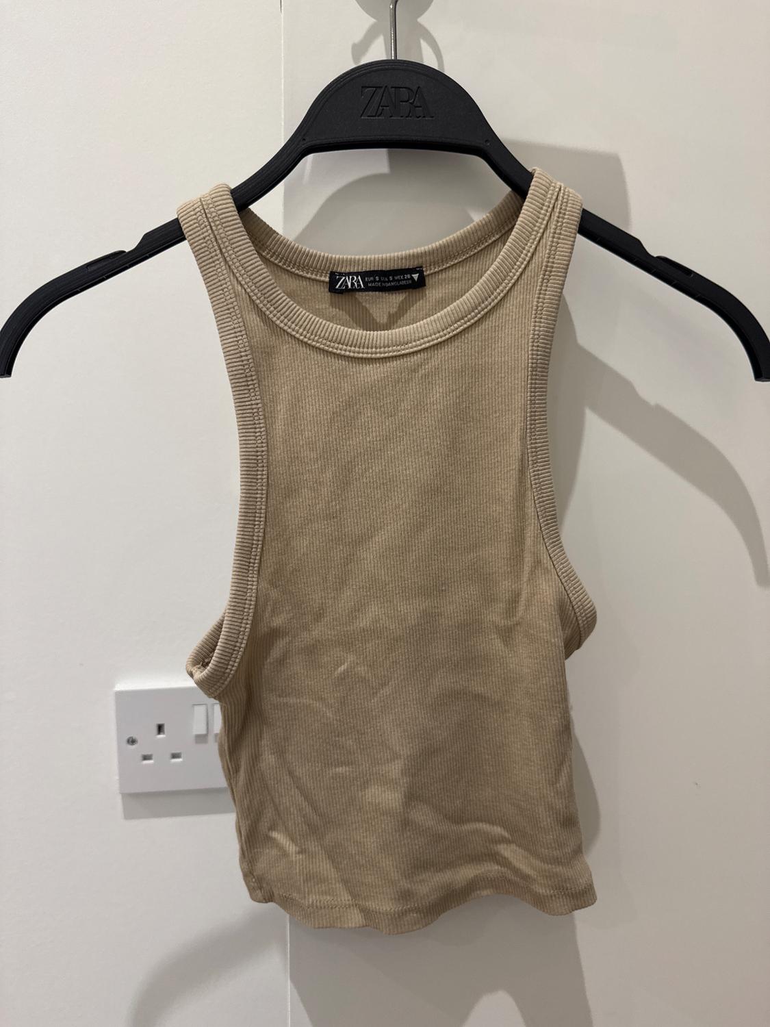 Beige sleeveless top hanging on a black hanger with the brand visible. White background with a plug socket on the wall.