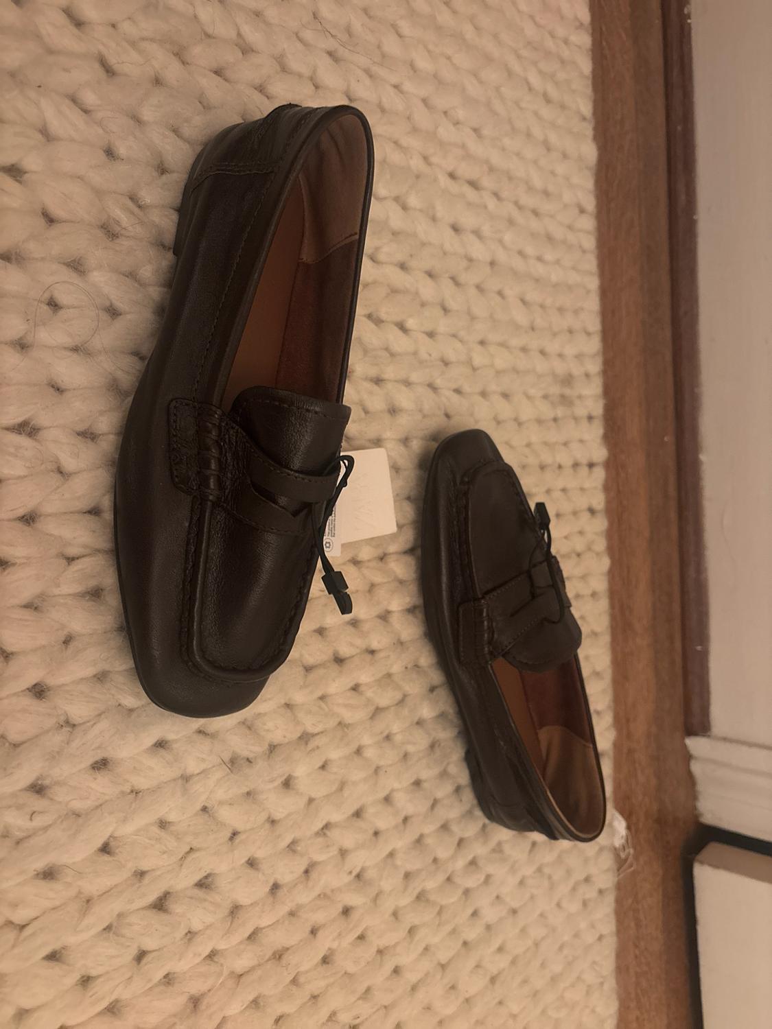 Pair of dark brown leather loafers with tags, on a cream-colored chunky knit rug next to a wooden floor.