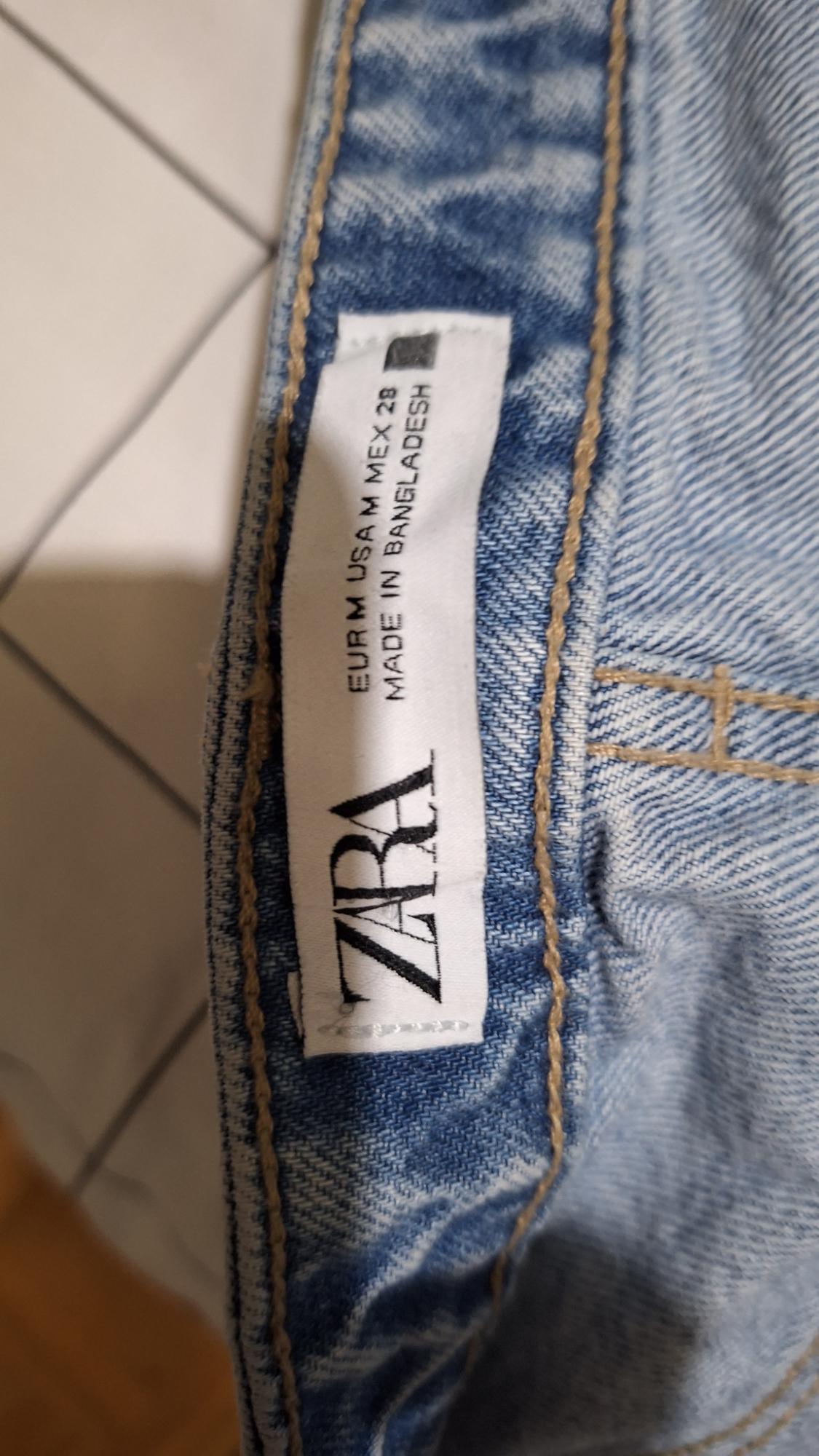 Label from blue jeans with visible stitching. Text: "ZARA EUR M USA M MEX 28 MADE IN BANGLADESH".