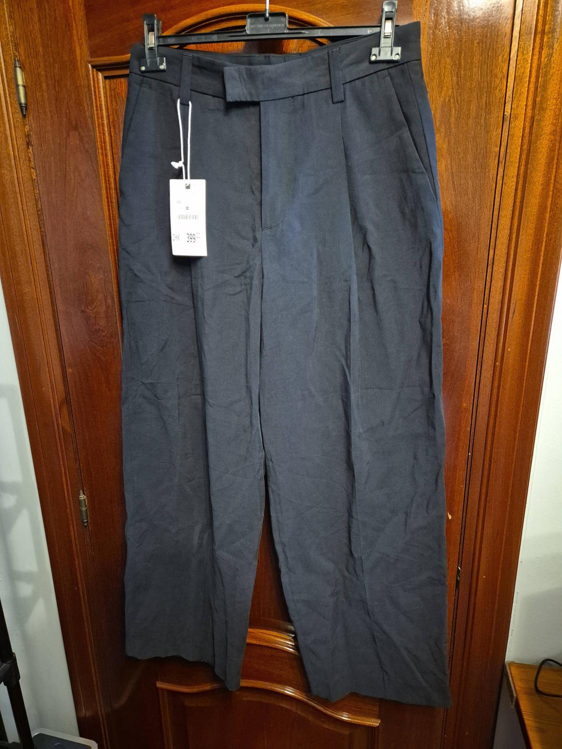 Dark grey wide-leg trousers hanging on a coat hanger over a wooden door. They have a tag bearing the text DKK 399.