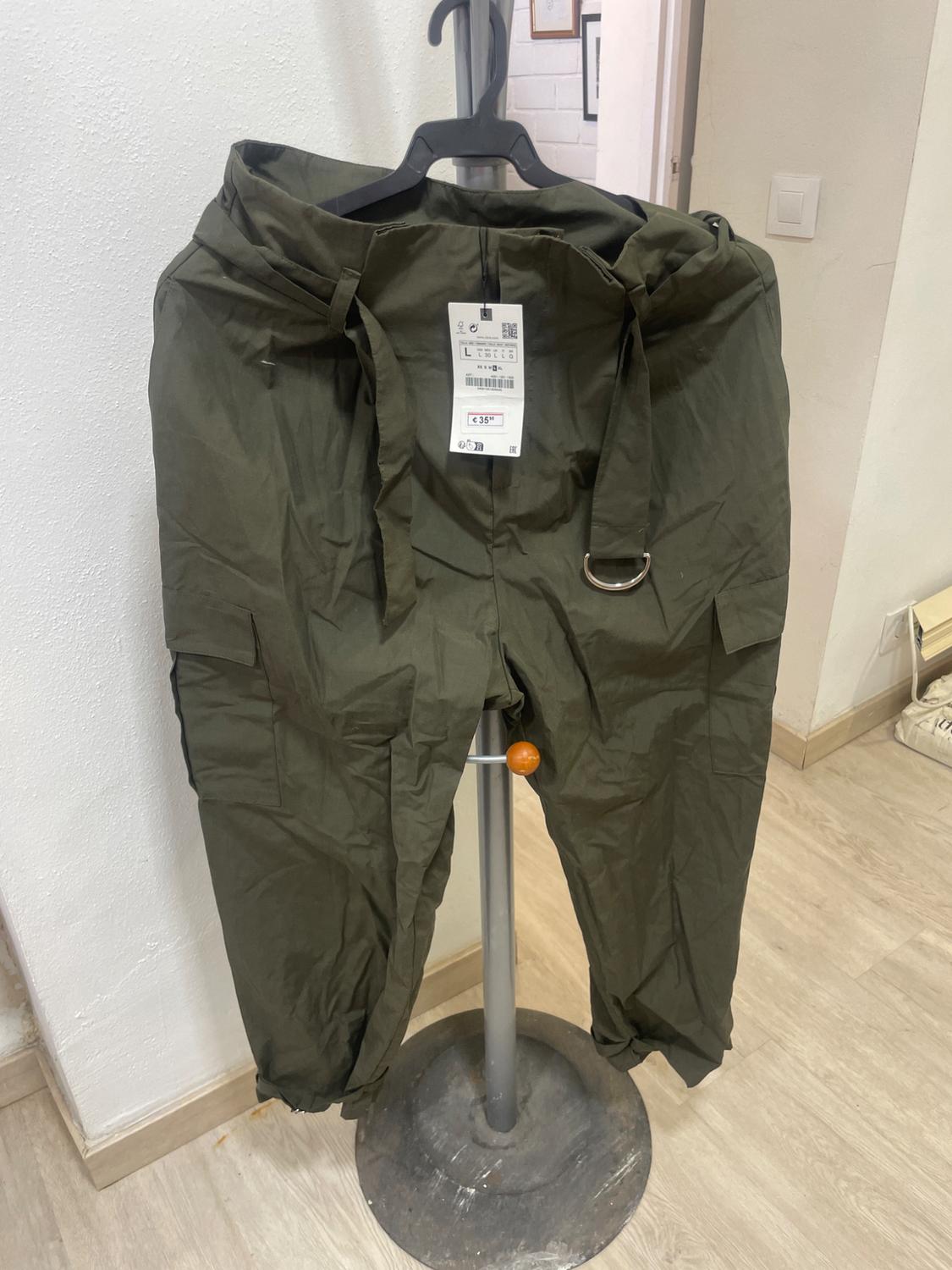 Green trousers hanging on a hanger with a price tag of £35. White wall background and light wood floor.