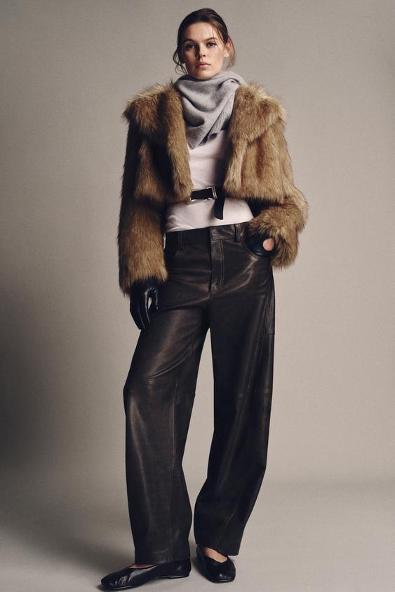 Komplet: krátká béžová bunda s chlupatým efektem, šedá šála, tmavé široké kalhoty a černé rukavice.