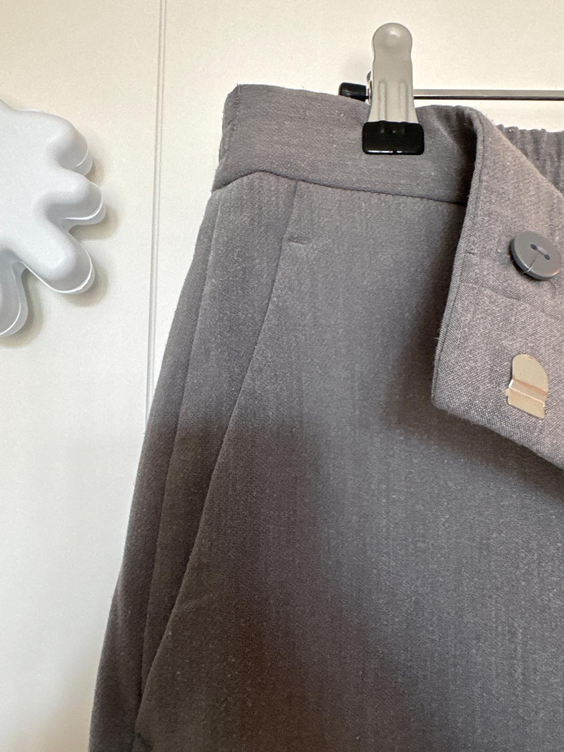 Grey trousers hanging on a hanger with black clips, featuring a button detail and elasticated waistband. White background with a cloud-shaped embellishment.