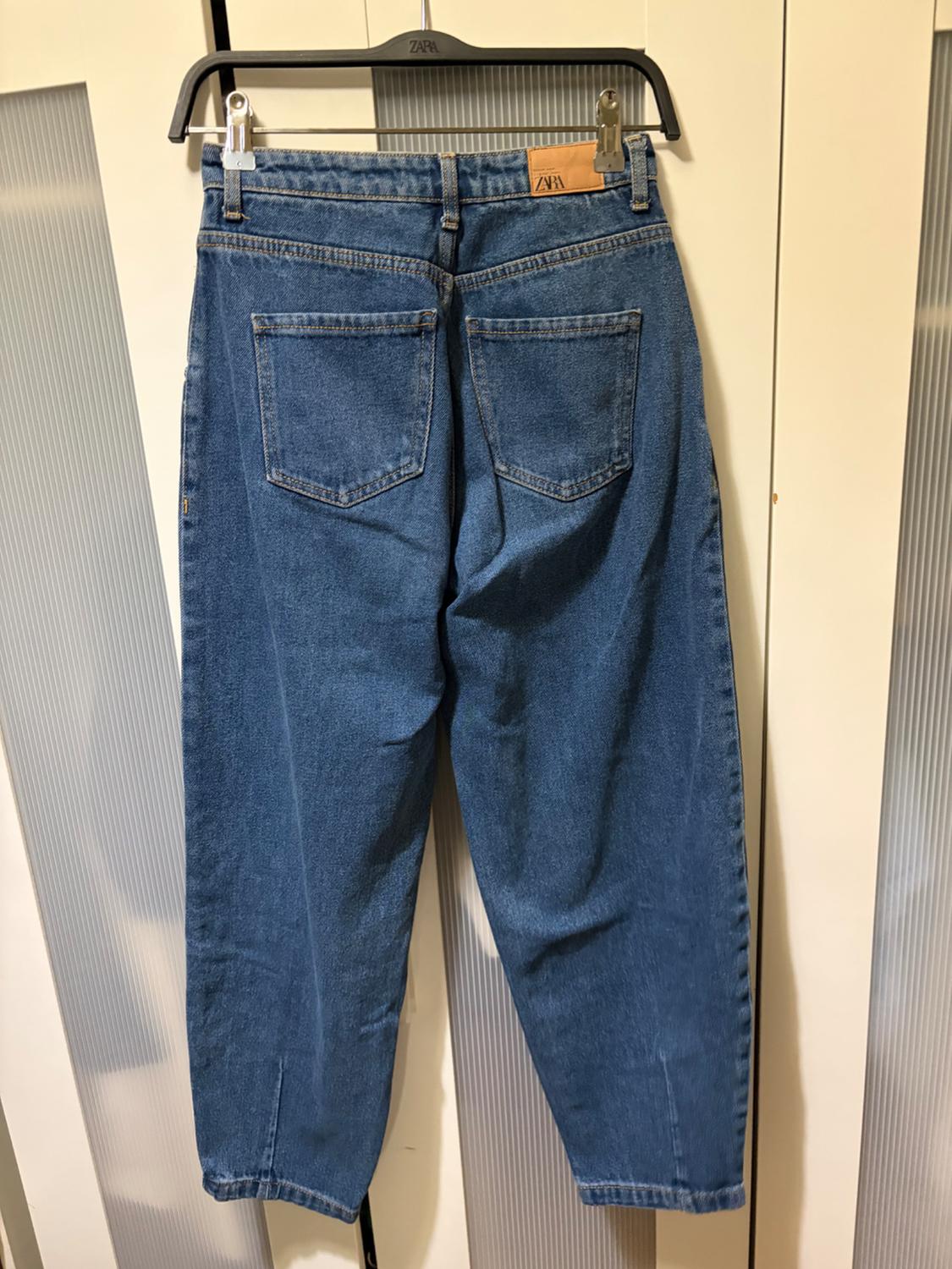 Blue jeans hanging on a black hanger with the "ZARA" label visible, against a background of white doors.