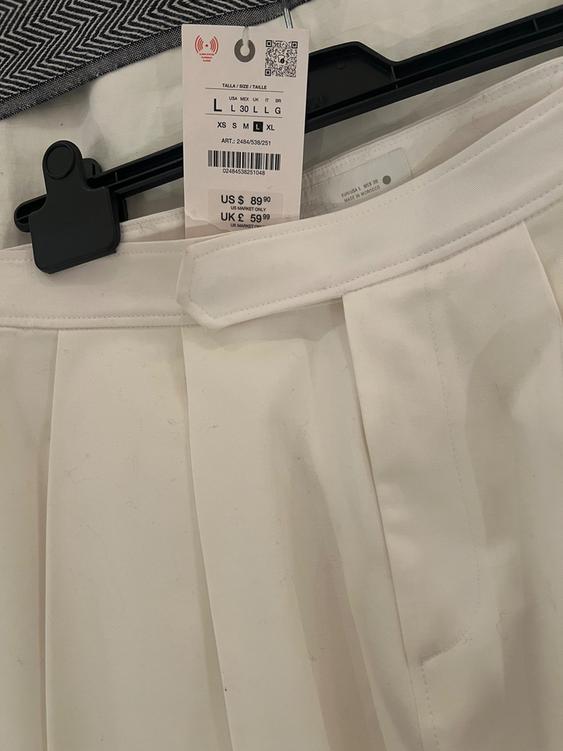 White pleated skirt tag on black hanger. Visible text: "TALLA/SIZE/TALLE L 30 L G XS M L XL US $89.90 UK £59.99".