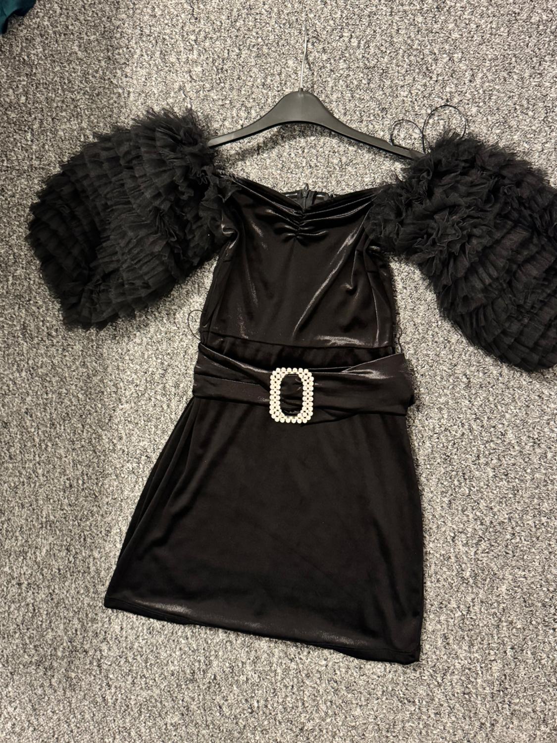 Black dress with voluminous pleated tulle sleeves, belt with decorative pearl buckle, hanging on a hanger over a grey rug.