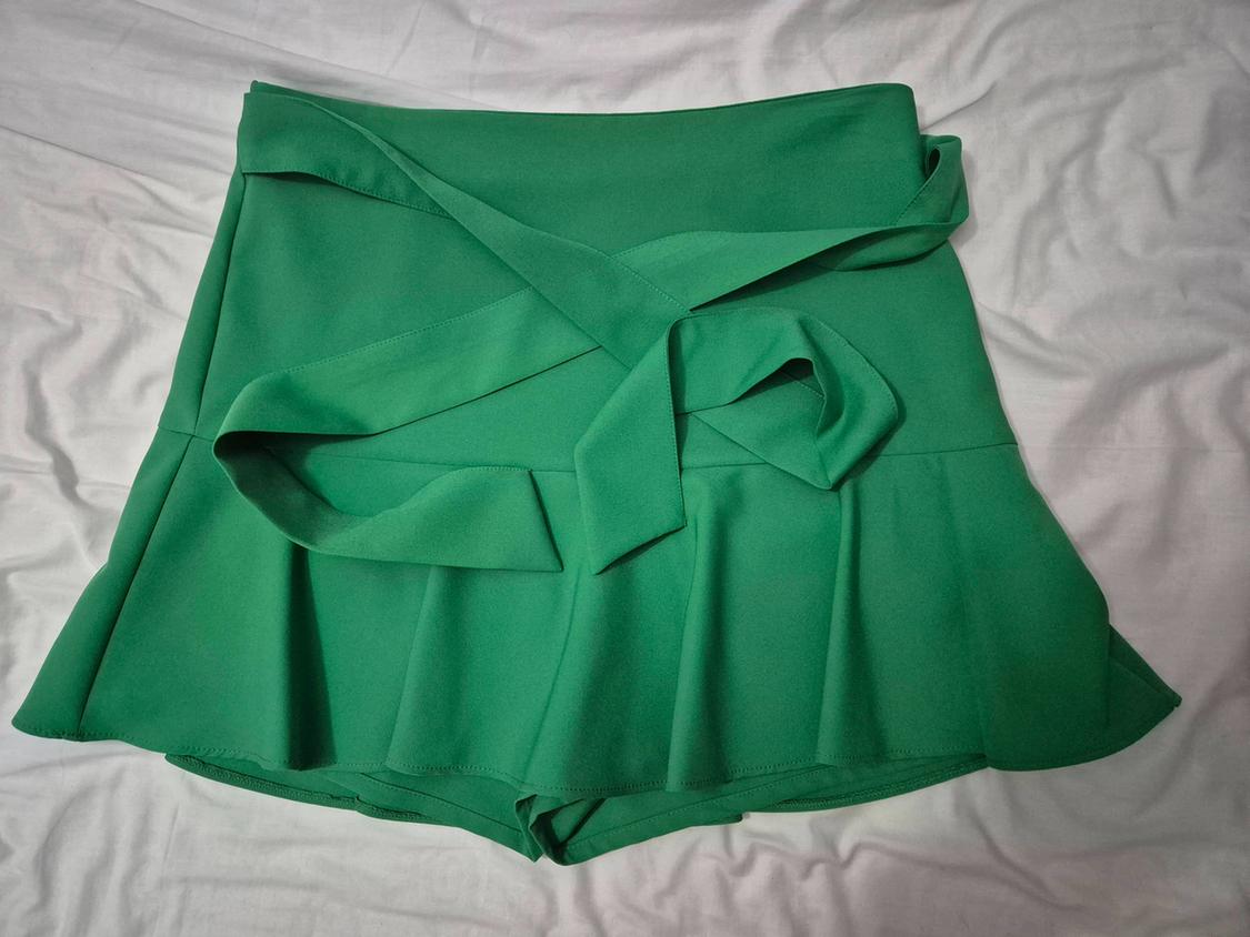 Green culottes with a tie-waist and flared hem, set against a crumpled white fabric that evokes a relaxed style.