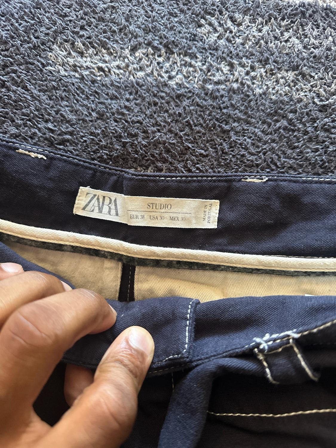 A hand holds a dark blue pair of pants, showing the label: ZARA STUDIO EUR 38 USA 30 MEX 30 MADE IN PORTUGAL. Gray carpet background.