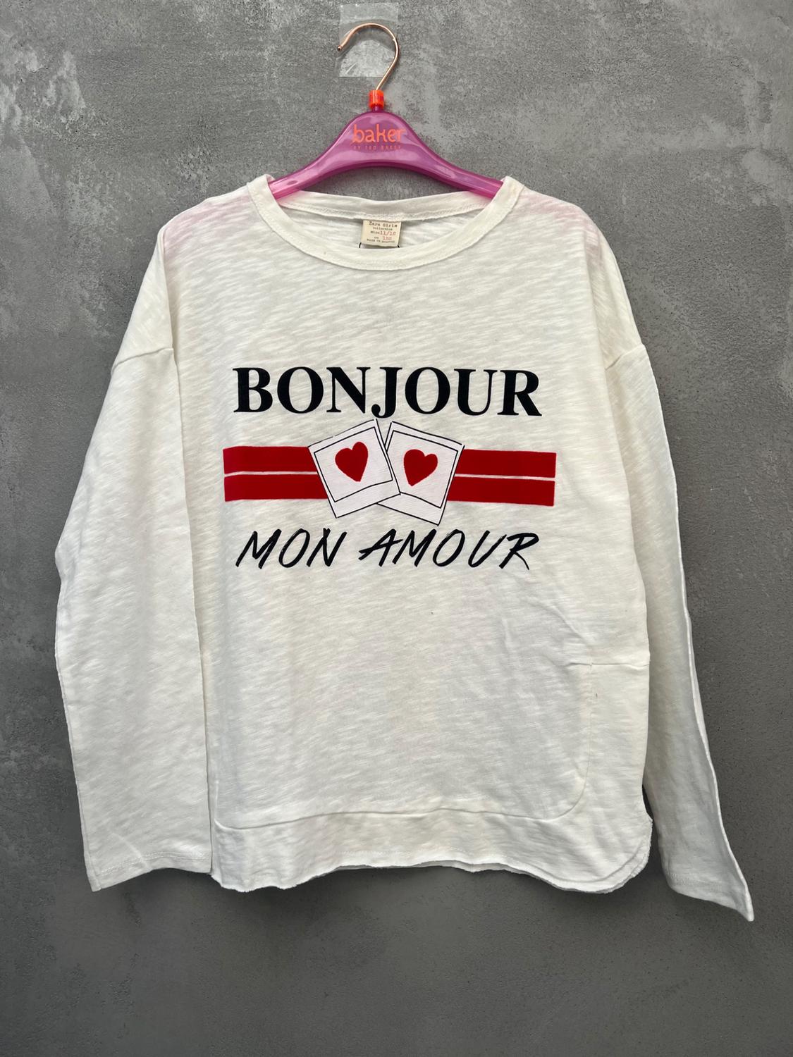White t-shirt with "BONJOUR MON AMOUR" text and two cards with red hearts, hanging on a pink hanger against a grey background.