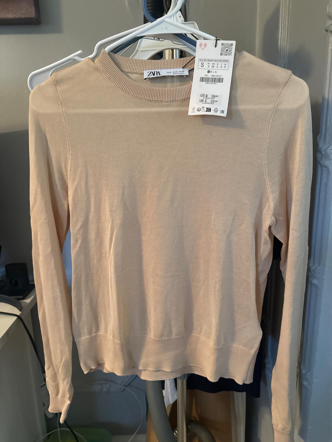 Beige long-sleeved jumper hanging on a white hanger. Visible tag with price in dollars and pounds. Indoor background.