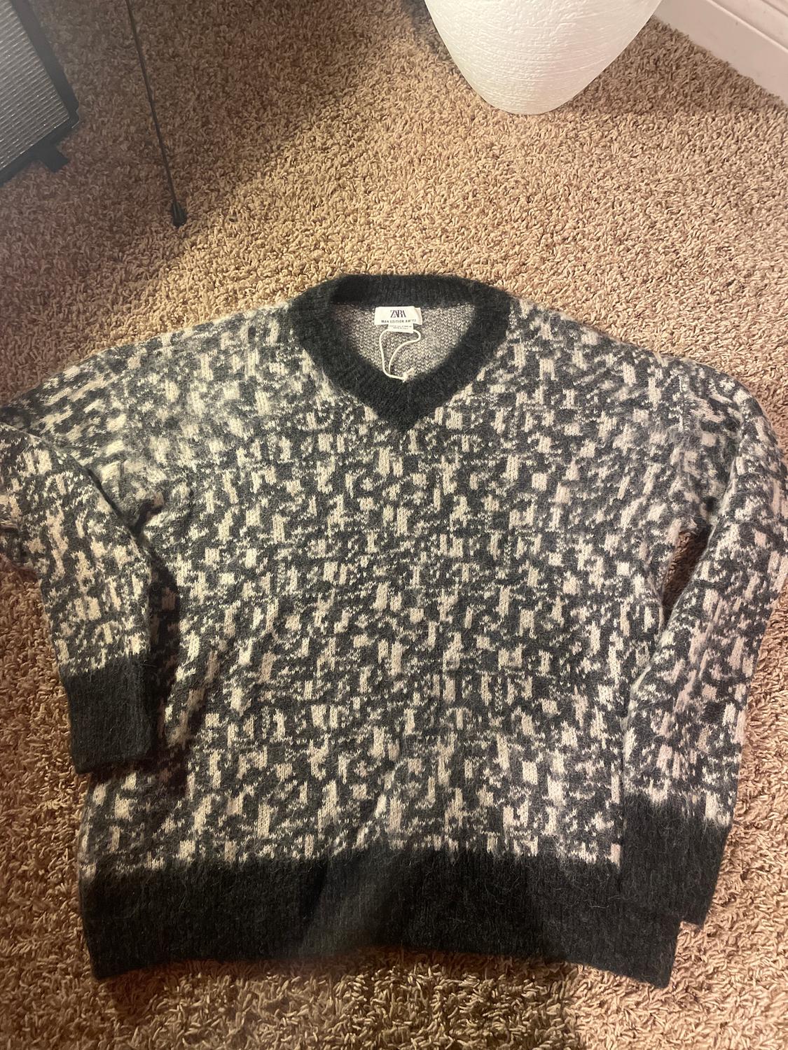 Grey and black knitted jumper with an abstract pattern, V-neck, and dark cuffs, on a beige rug. Visible label: "ZARA".