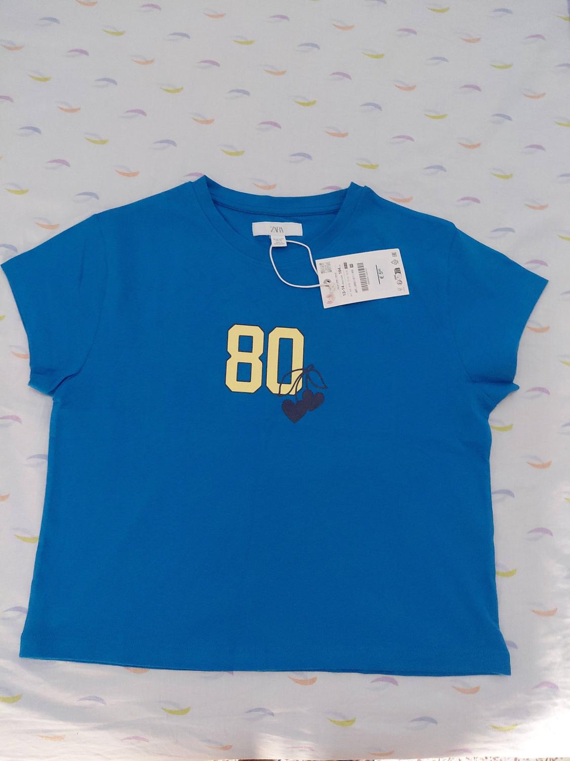 Blue short-sleeved T-shirt with the number 80 in yellow and some cherries. ZARA label visible. White background with a colourful print.