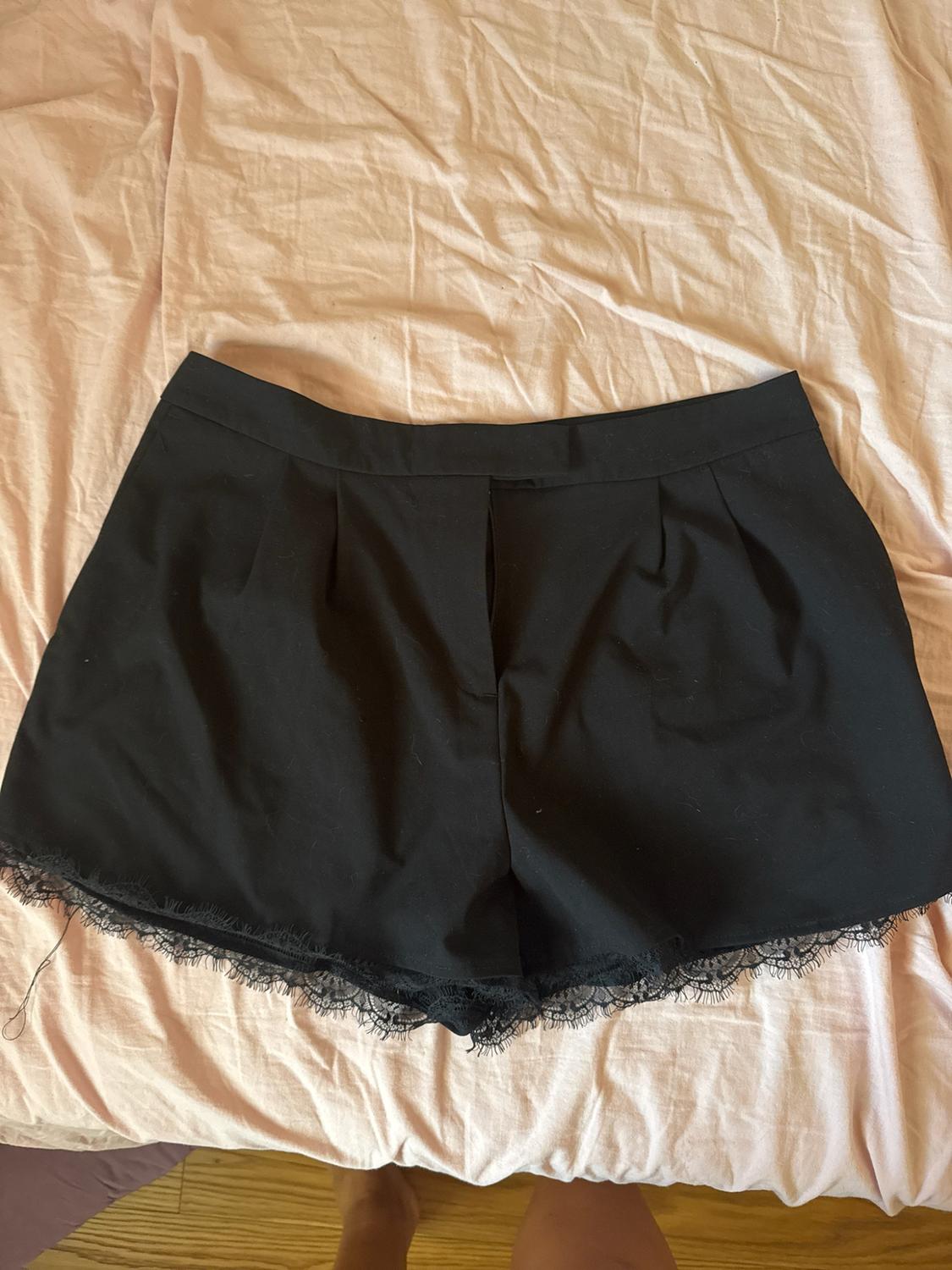 Black tailored shorts with lace trim, on a rumpled pink sheet. Casual style and relaxed atmosphere.