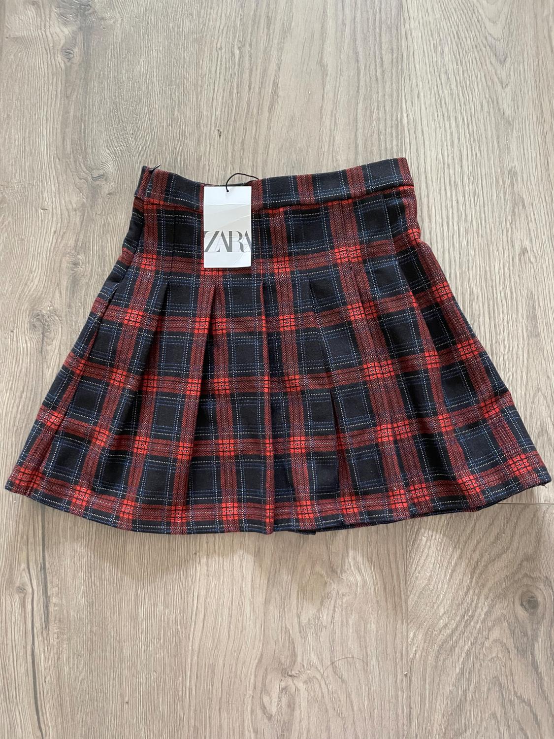Red and black plaid skirt with pleats on light wood flooring. White tag with "ZARA" text hanging.