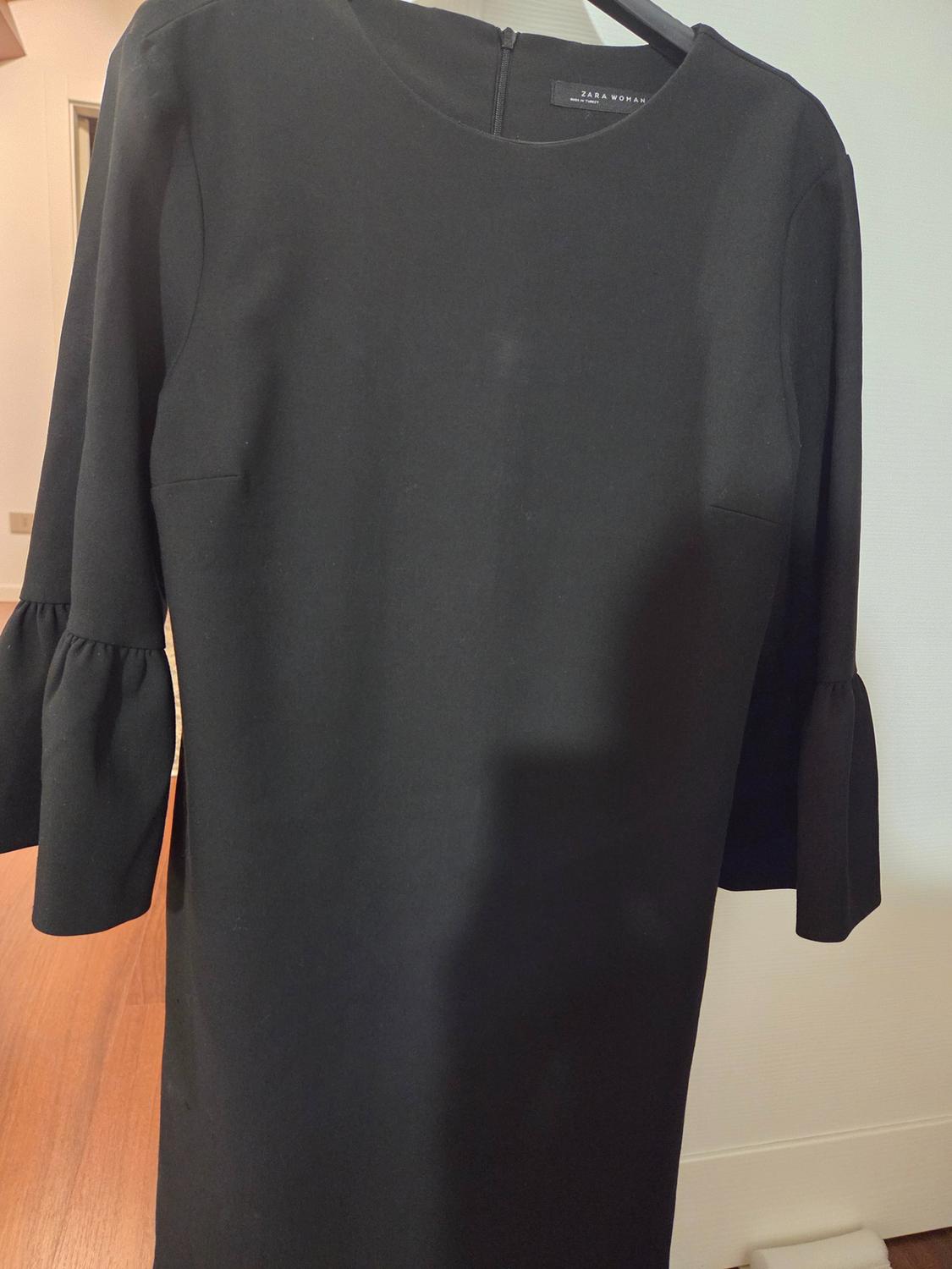 Black long-sleeved dress with ruffled cuffs, hanging on a hanger. Visible label: "Zara Woman". Indoor background.